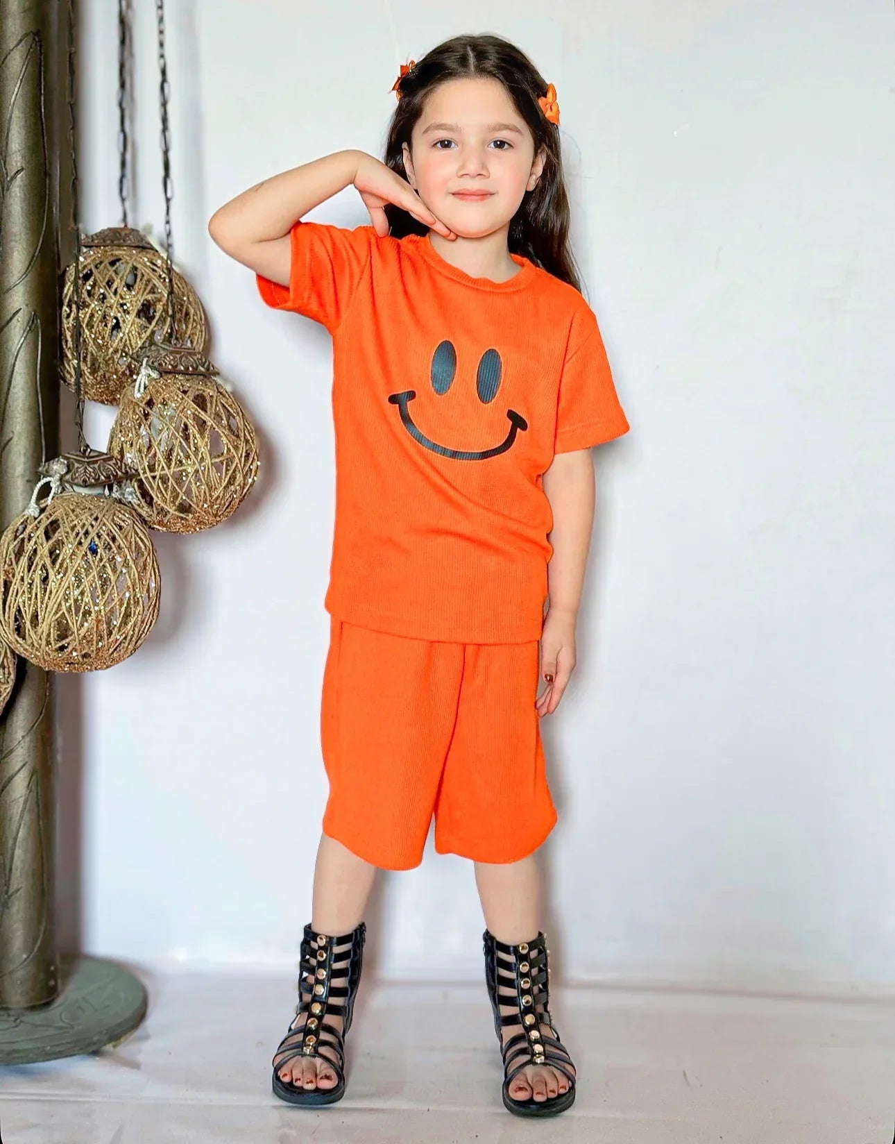 Child wearing an orange outfit with a smiley face design, standing against a white wall.