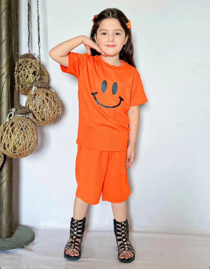 Child wearing an orange outfit with a smiley face design, standing against a white wall.