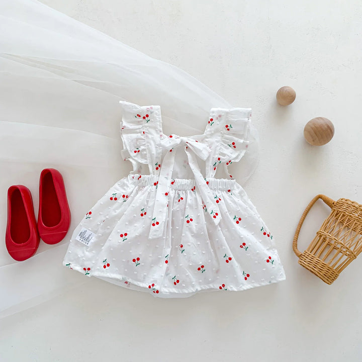 White dress with red cherry pattern on a white surface with red shoes and a wicker basket.
