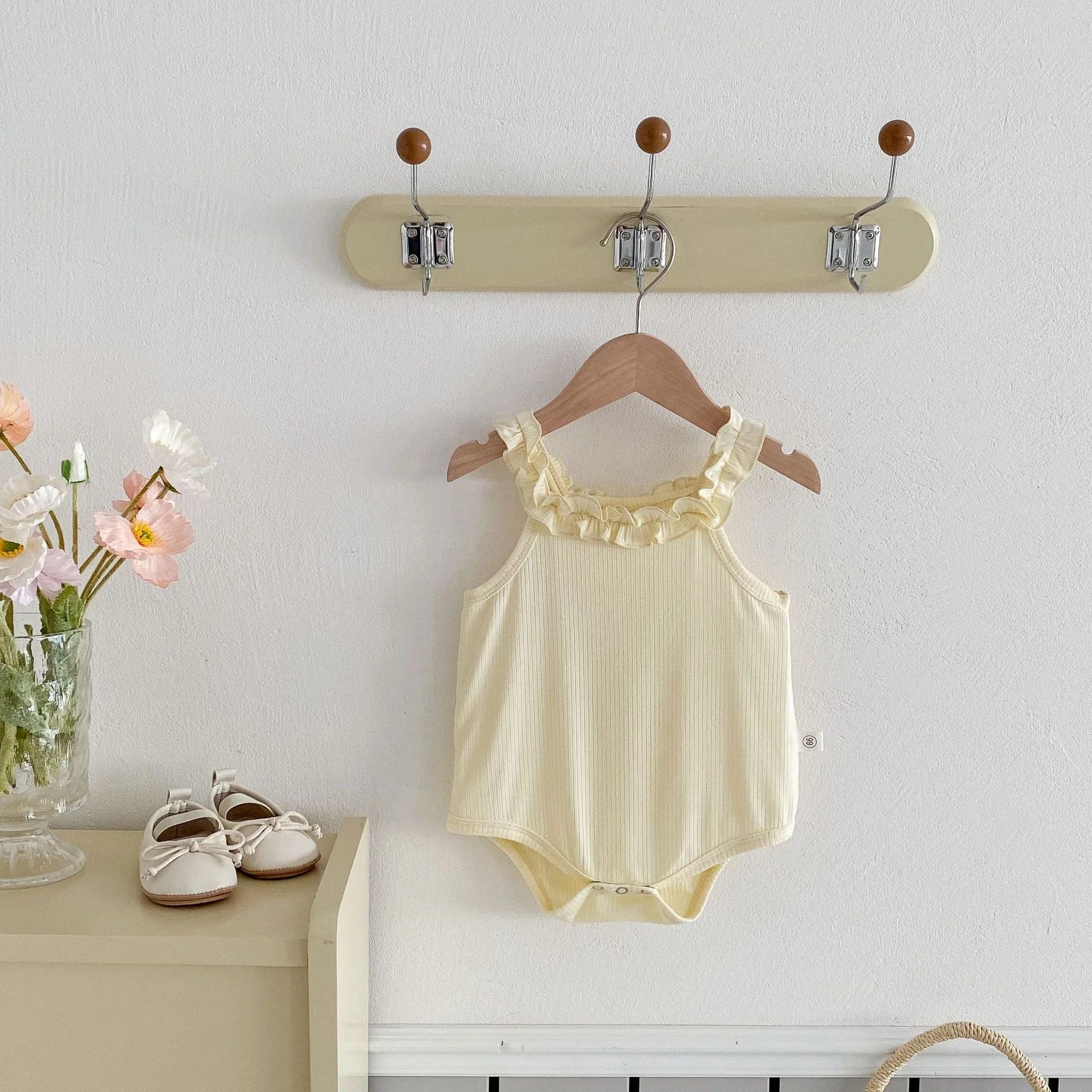 Yellow baby bodysuit on a hanger with a wall-mounted hook holder, flowers, and shoes in the background.