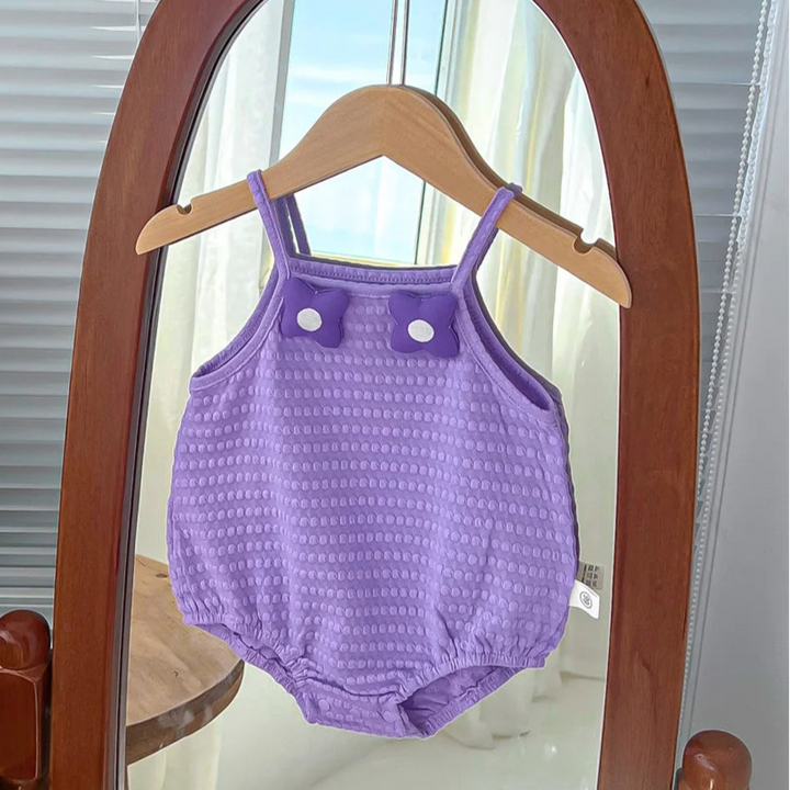 Purple baby romper hanging on a wooden hanger in front of a mirror with flowers and a box in the background.