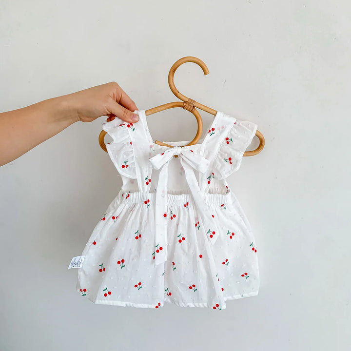 White baby dress with red cherry pattern on a wooden hanger against a light gray background
