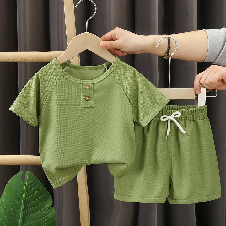 Green children's outfit with shirt and shorts on a hanger against a dark curtain background.