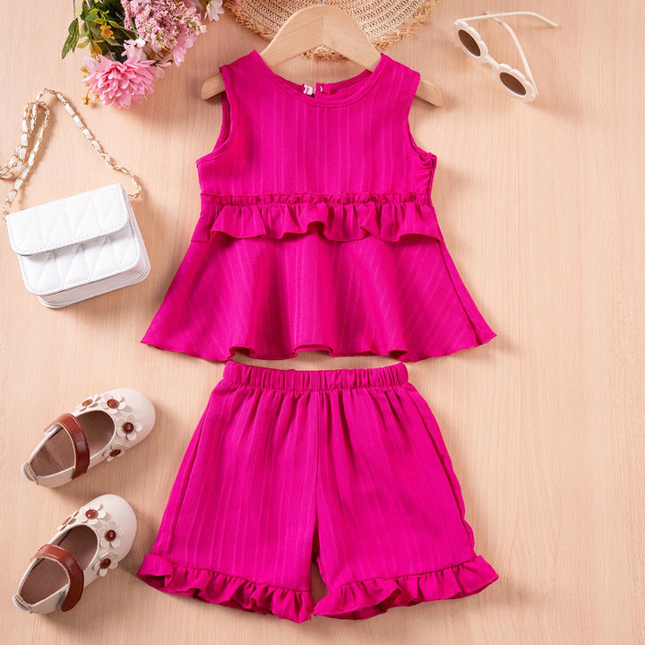 Pink children's outfit with top and shorts on a beige surface, surrounded by accessories.