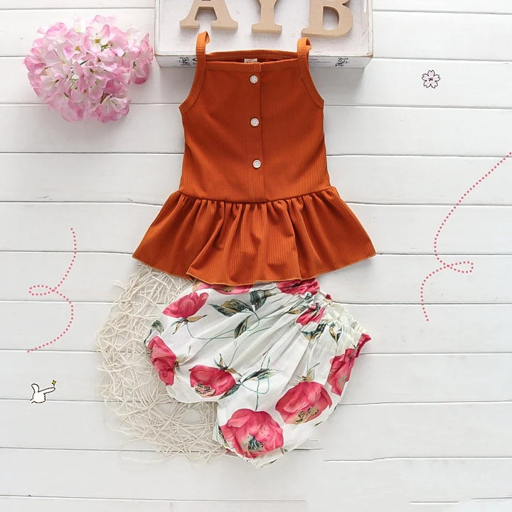Orange sleeveless top and floral bloomers on a white wooden surface with pink flowers and letters in the background.