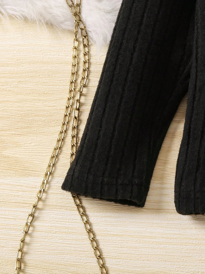 Black ribbed sweater sleeve with gold chain on a textured beige surface