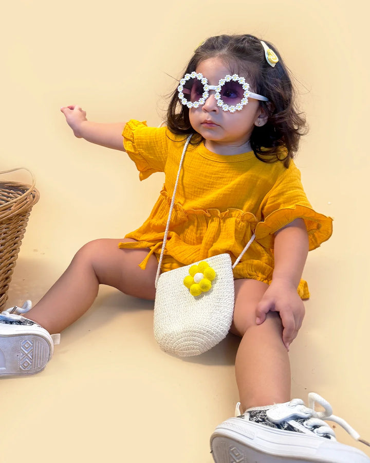 Child in a yellow outfit with sunglasses and a small white bag, sitting on a beige surface.