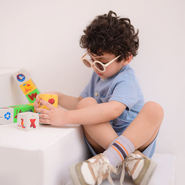 A young boy with curly hair wearing round sunglasses and a light blue outfit, sitting on white steps while playing with colorful alphabet blocks.