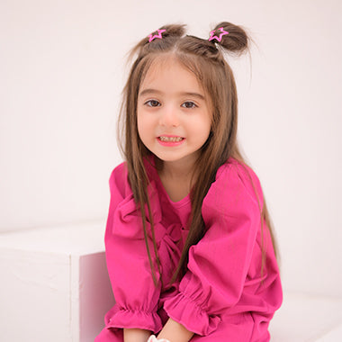 A smiling young girl with long brown hair styled in two space buns with pink star clips, wearing a bright pink outfit, sitting indoors against a light background.