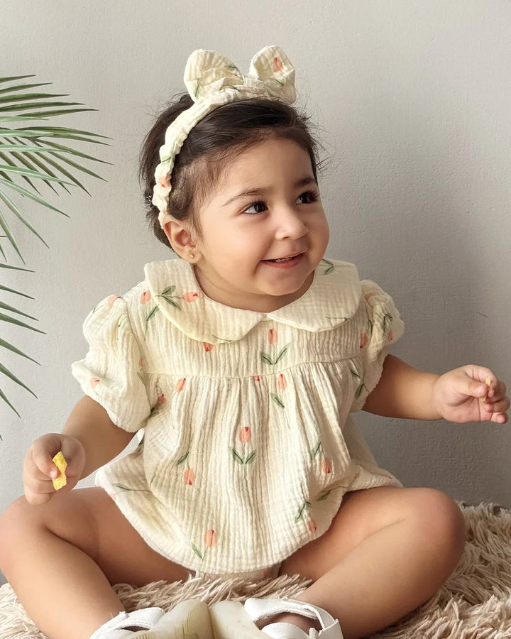 Baby wearing a floral romper with a headband, sitting on a soft surface.
