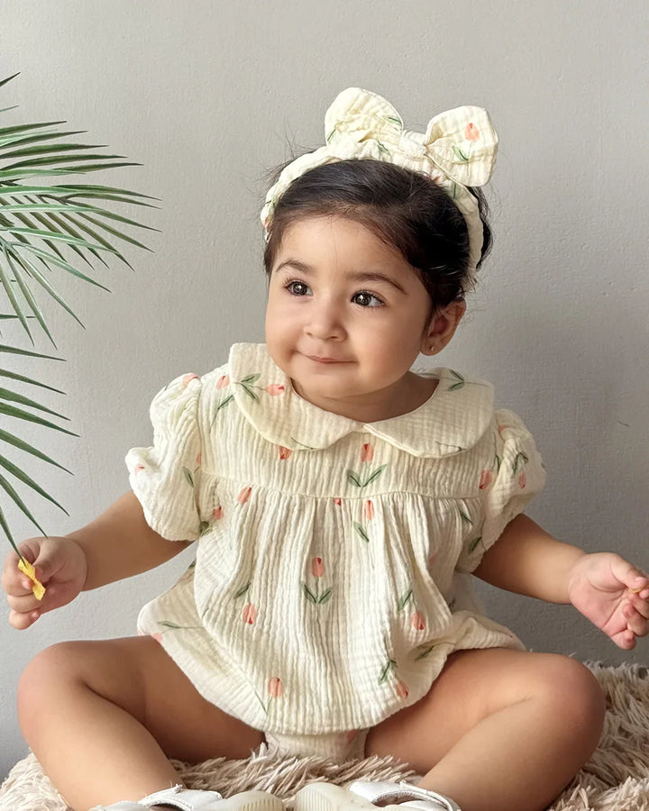 Baby wearing a floral outfit with a headband, sitting on a textured surface with a plant in the background.