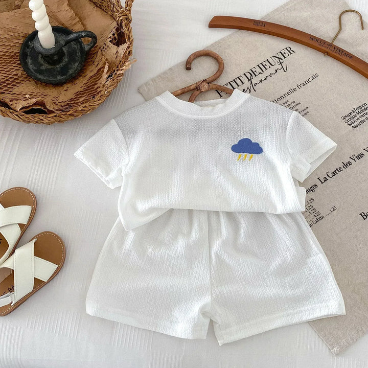 White baby outfit with cloud design on shirt, placed on a textured surface with a hanger and basket in the background.