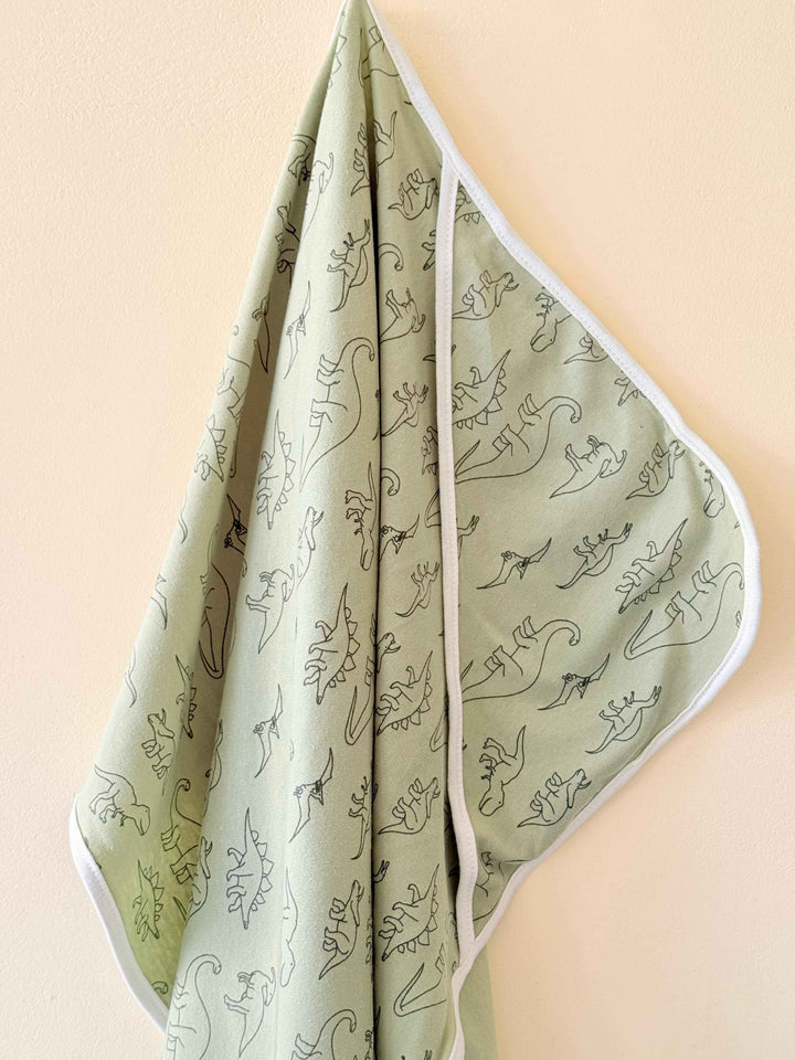 Sage green baby blanket with black dinosaur illustrations and white trim hanging against a beige wall.