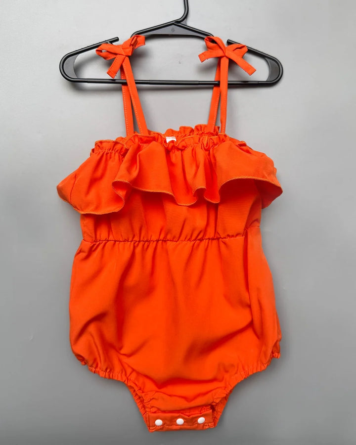 Orange baby romper with ruffled top and bow straps on a hanger against a gray background