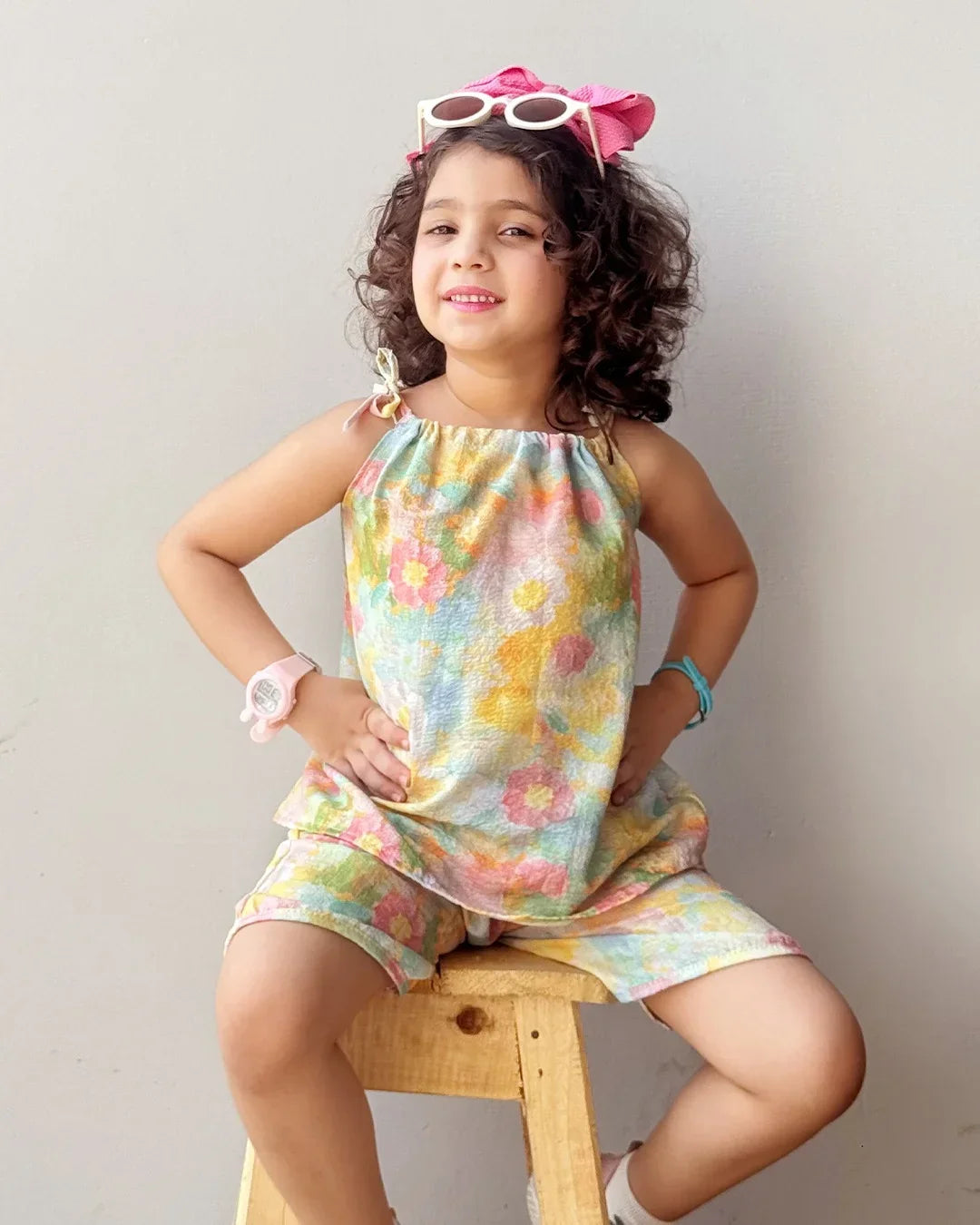 Young girl wearing a colorful floral dress sitting on a wooden stool against a plain background