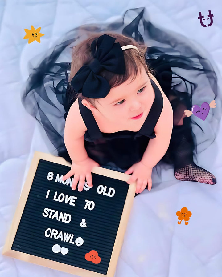 Baby in a black dress holding a sign saying '8 months old, I love to stand & crawl' on a white blanket.