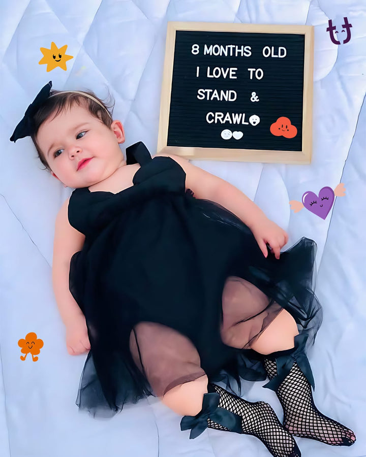 Baby in a black outfit with fishnet tights and a sign indicating 8 months old, standing and crawling.