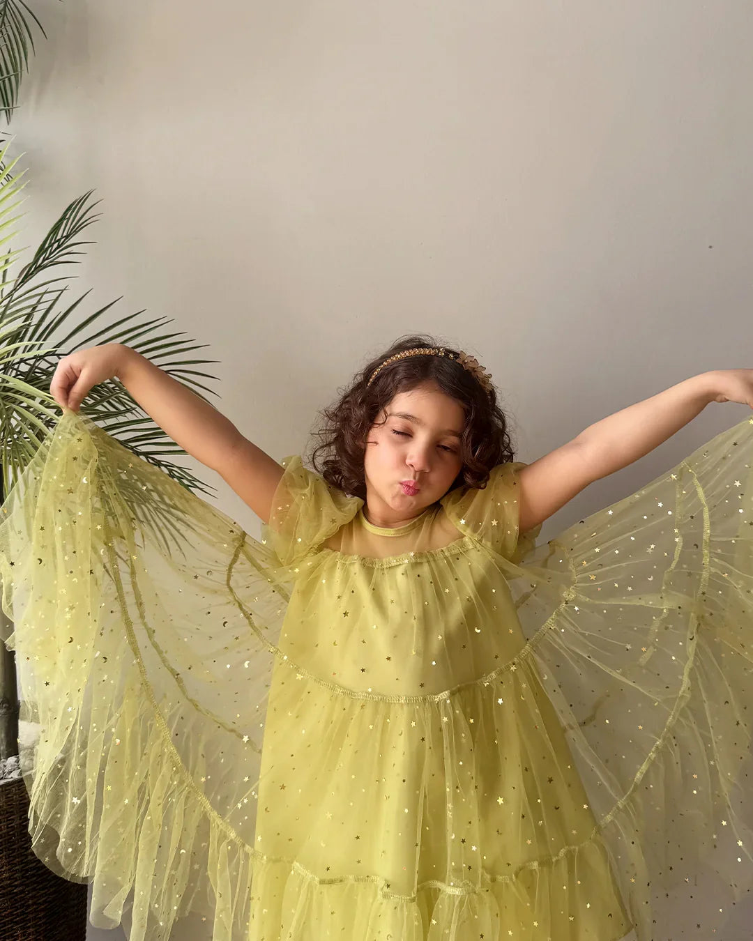 Child in a yellow fairy costume with wings standing next to a plant indoors.