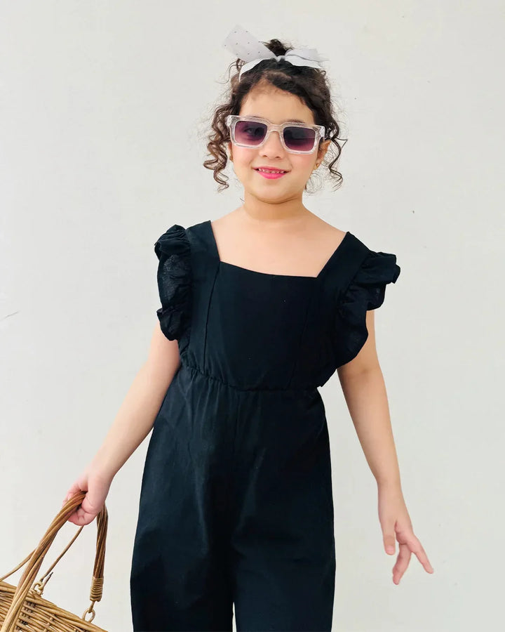 Young girl wearing a black ruffled jumpsuit with sunglasses and a white headband against a plain background