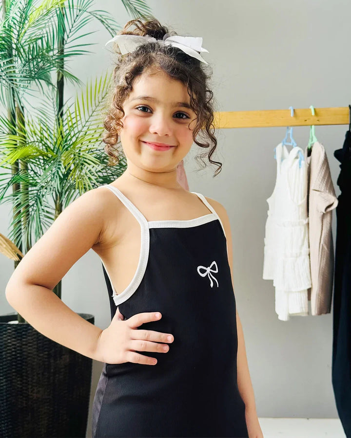 Young girl wearing a black tank top with a white bow design, standing in a room with plants and clothing racks.