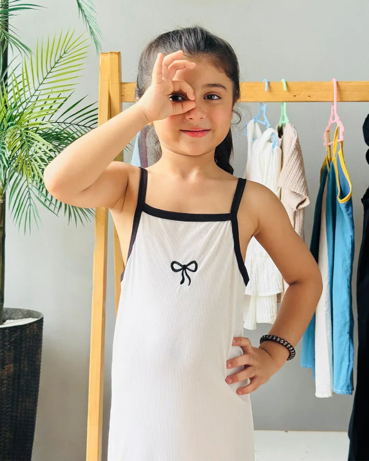 Child wearing a white dress with a black bow design, standing in front of a rack of clothes.