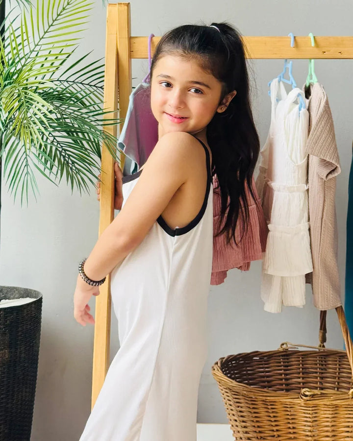 Young girl in a white dress standing in front of a rack with clothes, next to a plant and wicker basket.