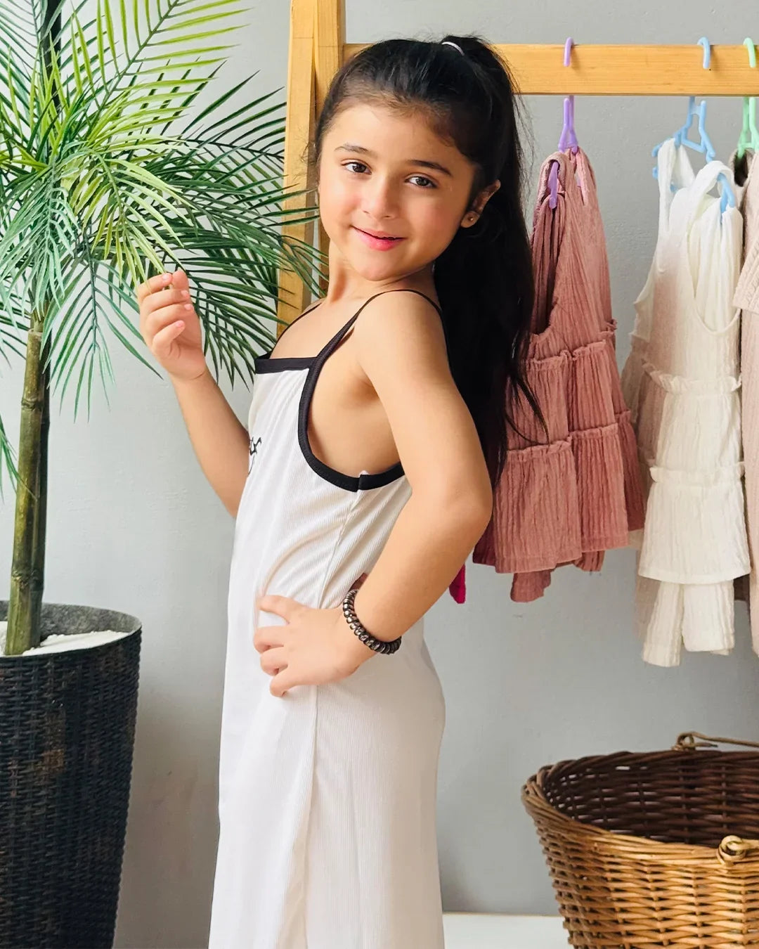 Young girl in a white dress standing in front of a rack of clothes with a plant and basket in the background.