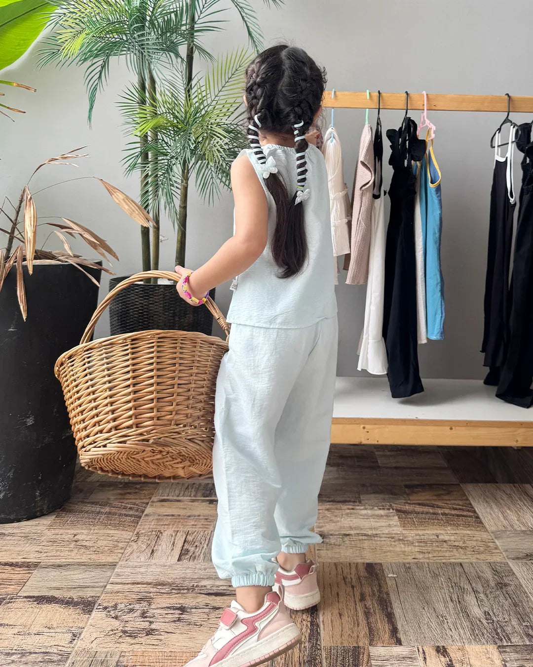 Child in light blue outfit holding a basket, standing in front of a clothing rack with various garments.
