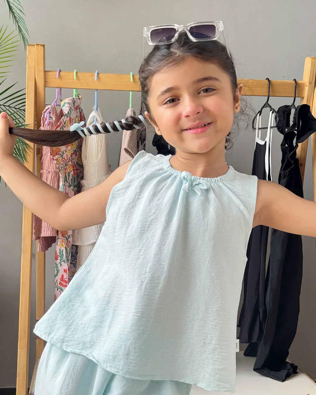 Young girl in a light blue dress holding a black belt with a rack of clothes in the background