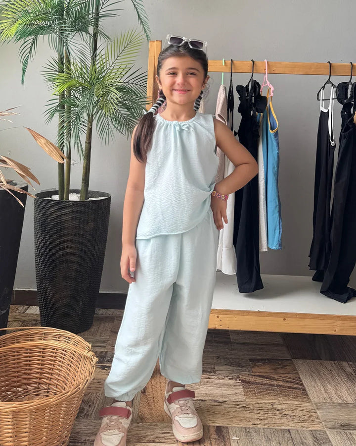 Young girl in light blue outfit standing in a room with plants and clothing racks.