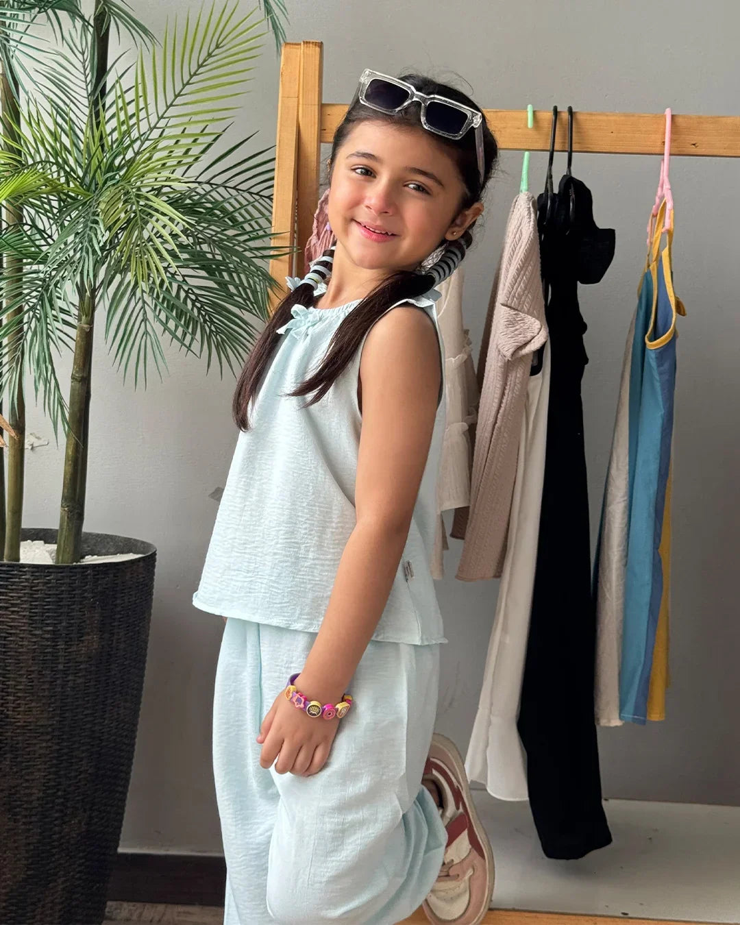 Young girl in light blue outfit standing in front of a rack with clothes, next to a plant.