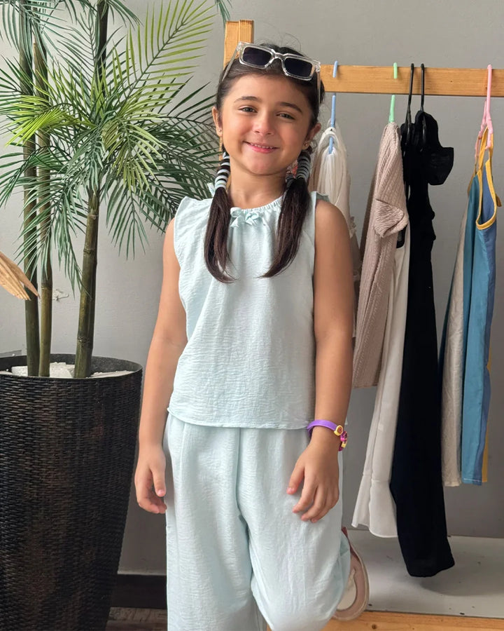 Young girl in light blue sleeveless top and pants standing in front of a clothing rack with plants in the background.