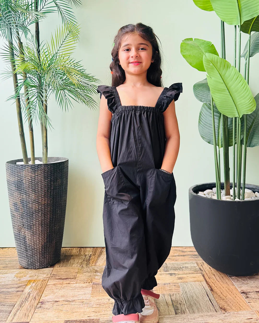 Young girl in a black dress standing between two potted plants indoors.