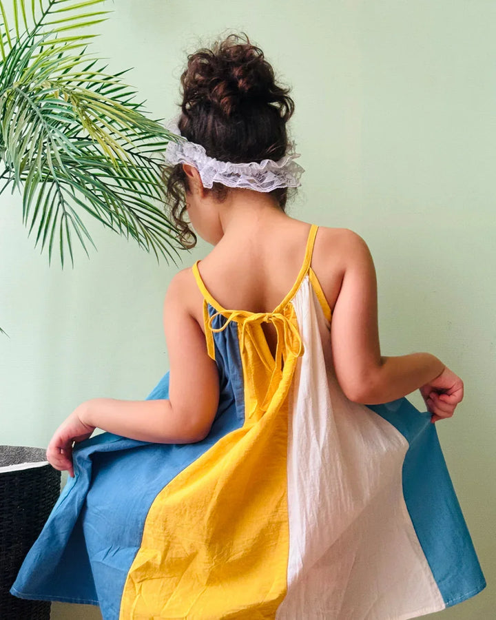 Child wearing a yellow and blue dress with a white headband, sitting on a chair against a light green wall.
