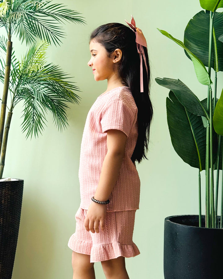 Young girl in a pink outfit standing between two potted plants with a light green wall background
