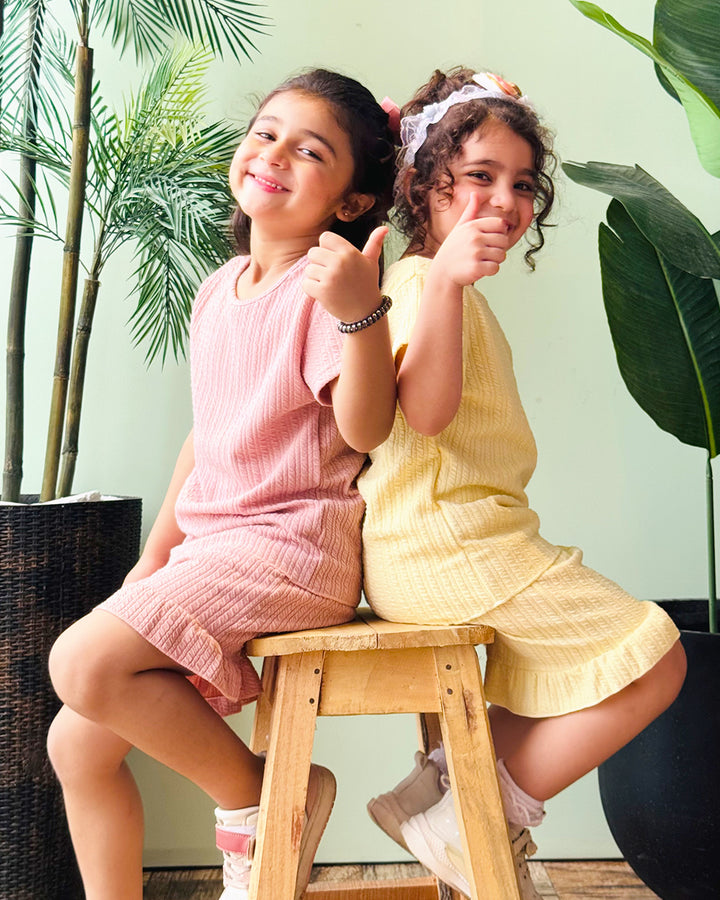 Two young girls sitting on a wooden stool, giving thumbs up, with plants in the background.