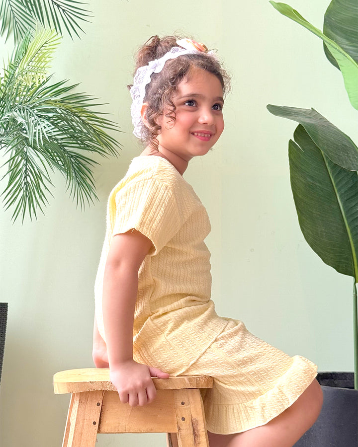Young girl in a yellow outfit sitting on a wooden stool with plants in the background