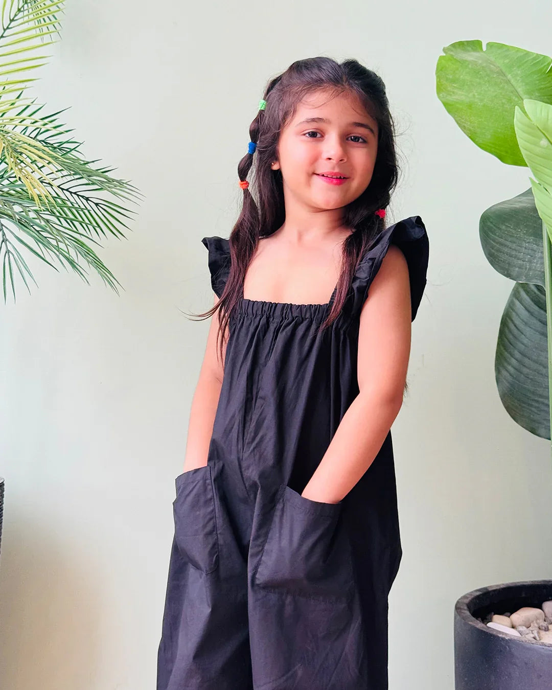 Young girl wearing a black dress with ruffled sleeves against a light green wall with plants.