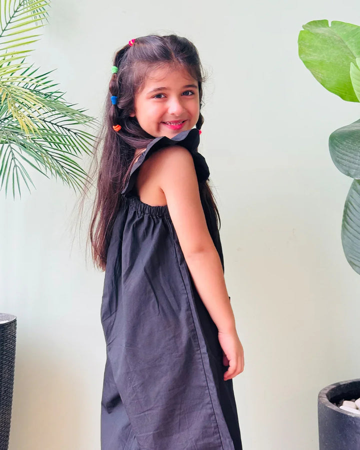 Young girl wearing a black sleeveless dress with colorful hair accessories, standing against a light background with green plants.